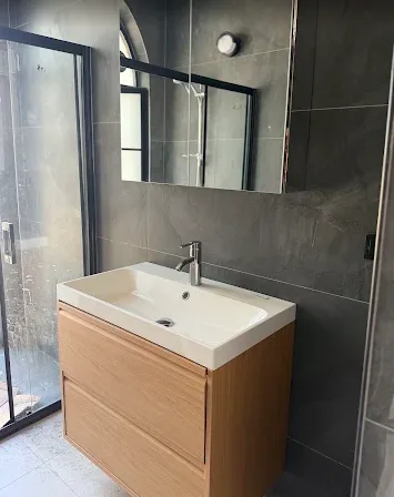Yenilenmiş banyoda ahşap dolaplı lavabo, aynalı dolap ve siyah çerçeveli duşakabin